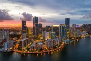 Aerial view of a coastal cityscape with high-rise buildings illuminated at dusk. The sky displays a mix of clouds and warm colors from the setting sun, reflecting on the water. Streets and buildings along the waterfront are lit up, creating a picturesque scene.