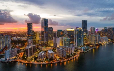 Aerial view of a coastal cityscape with high-rise buildings illuminated at dusk. The sky displays a mix of clouds and warm colors from the setting sun, reflecting on the water. Streets and buildings along the waterfront are lit up, creating a picturesque scene.