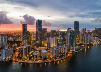 Aerial view of a coastal cityscape with high-rise buildings illuminated at dusk. The sky displays a mix of clouds and warm colors from the setting sun, reflecting on the water. Streets and buildings along the waterfront are lit up, creating a picturesque scene.