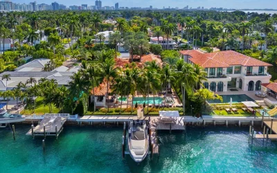 Aerial view of a luxury waterfront mansion with a pool, surrounded by palm trees, near a docked yacht in a tropical setting. Nearby city skyline in the background. Edward Blackburn, Licensed Investment Counselor. Your source for North Georgia real estate.