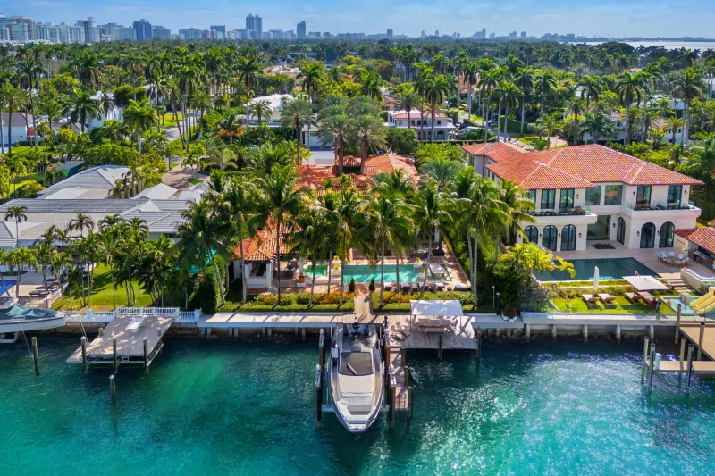 Aerial view of a luxury waterfront mansion with a pool, surrounded by palm trees, near a docked yacht in a tropical setting. Nearby city skyline in the background. Edward Blackburn, Licensed Investment Counselor. Your source for North Georgia real estate.